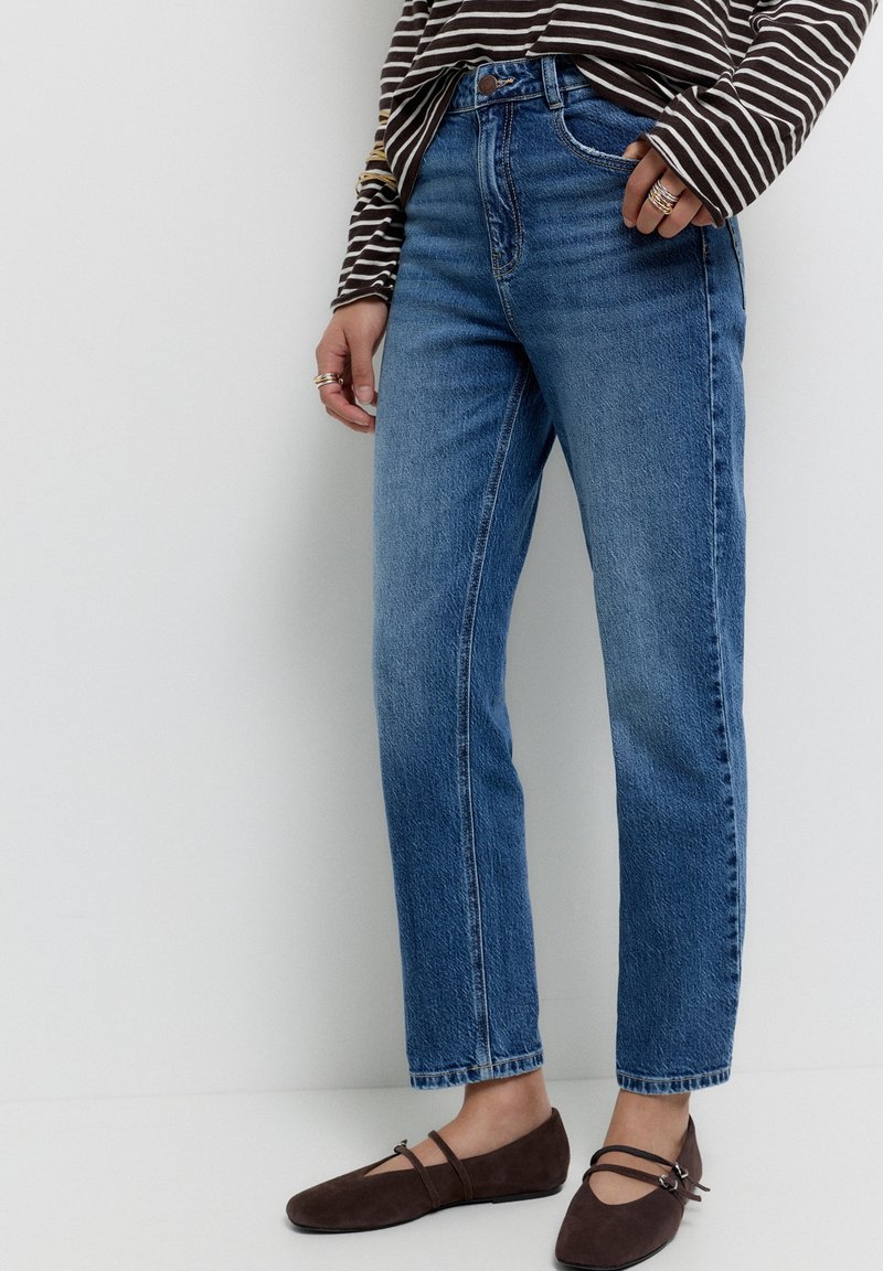 Femme portant un jean bleu taille haute à jambes droites, un haut à manches longues rayé, des ballerines Mary Jane en daim marron et plusieurs bagues aux doigts.