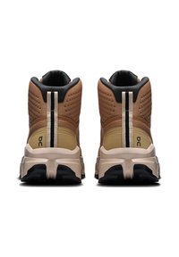 Pantofi sport înalți cu partea superioară din plasă maro, accente negre, talpă căptușită de culoare crem și un logo proeminent pe lateral cu un detaliu cu linie albă.