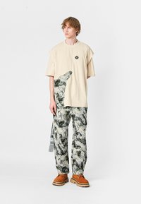 Joven con camiseta oversized beige con estampado abstracto en blanco y negro, pantalones a juego con patrón y zapatos marrones con cordones, de pie.