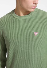 Maglione verde lavorato a maglia con texture a coste, scollatura rotonda e un piccolo logo triangolare rosso e bianco sul lato sinistro del petto.