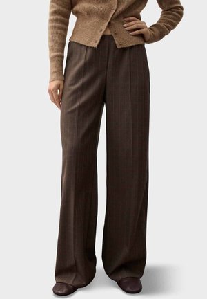 Femme portant un pantalon large à fines rayures marron, un cardigan côtelé beige et des chaussures marron foncé, debout, une main sur la hanche.