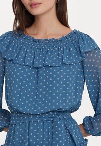 Kvinna i en blå långärmad klänning med vita prickar, volangkrage och matchande tygbälte knutet i midjan.