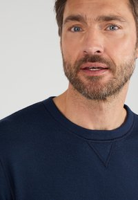 Navy blauwe gebreide sweatshirt met een ronde hals, ribbelstructuur en zichtbare stiksels. Close-up beeld dat zich richt op de halslijn en schouder.