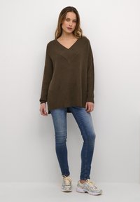Brun ribbad oversized tröja med djup V-ringning, kombinerad med blå smala jeans och ljusa träningsskor. Slät textur, avslappnad passform.