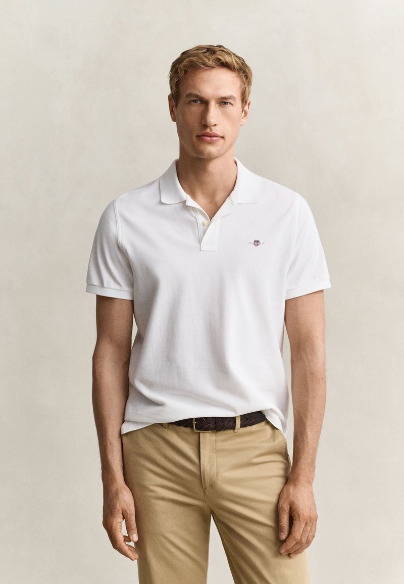 Jeune homme portant un polo blanc à manches courtes et un pantalon beige, debout devant un fond clair uni.