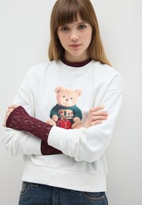 Jonge vrouw met lichtbruin haar, gekleed in een witte sweatshirt met een teddybeer die een cadeau vasthoudt en bordeauxrode kanten mouwen, staande tegen een witte muur.
