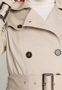 Béžový trenčkot s dvouřadým zapínáním, velkými knoflíky, páskem se čtvercovou sponou a texturovaným materiálem.