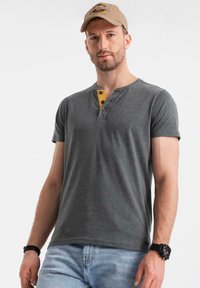 Krátký rukáv, tmavě šedé tričko henley s žlutým akcentem a knoflíky. Hladká tkanina. V kombinaci se světle modrými džíny a béžovou čepicí.