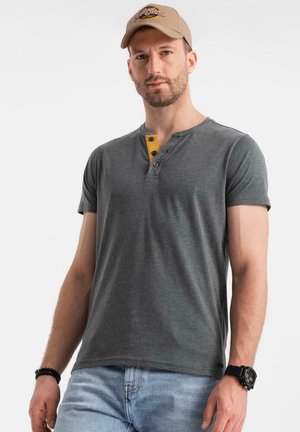 Camisa henley de manga corta en gris oscuro con acento amarillo y botones. Tejido suave. Combinada con jeans azul claro y una gorra beige.