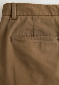 Pruunid puuvillased püksid, millel on sile tekstuur, tagatasku ja õmblustega detailid vöökoha ning tasku piirkonnas.