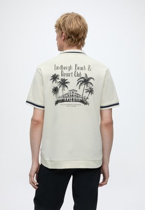 Blonder Mann, der von hinten zu sehen ist, trägt ein cremefarbenes Poloshirt mit schwarzer Palme, Resort-Gebäudegrafik und dem Schriftzug "Lindbergh Beach & Resort Club" auf dem Rücken.