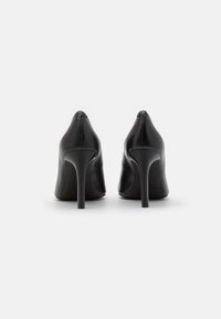 Un paio di scarpe nere in pelle con tacco alto viste da dietro su uno sfondo bianco.