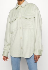 Giacca camicia in pile verde chiaro con colletto classico, due tasche sul petto e chiusure a bottoni. Texture morbida e vestibilità comoda.