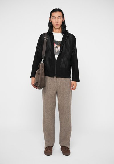 Juodas raukšlėtas užtrauktuko džemperis už baltos grafikos marškinėlių, smėlio spalvos tekstūruoti plačiakulkšniai kelnes ir rudos spalvos batus, su rudų rankinio krepšiu.