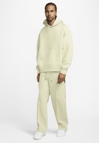 Moški z muze z litimi lasmi, oblečen v kremo pulover, ujemajoče se ohlapne hlače in bele superge, stoji ob belem ozadju.