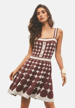 Žena nosi šaty bez rukávov s hnedými kruhovými vzormi a tromi gombíkmi na hrudi, doplnené zlatými náušnicami v tvare kruhu a širokým náramkom.