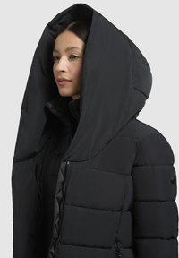 Giubbotto imbottito nero con un grande cappuccio unico. Realizzato in materiale leggero, presenta ampie sezioni trapuntate e una chiusura frontale con zip.