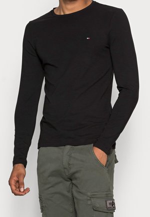 Chemise noire à manches longues en coton. Présente un petit logo sur la poitrine, un col rond et une coupe ajustée. Associée à un pantalon cargo olive.