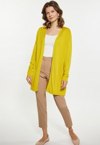 Lange, open voorkant cardigan in levendig geel, gemaakt van zachte stof. Draag over een witte top en gecombineerd met beige broeken. Eenvoudig ontwerp.