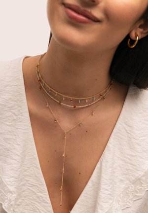 Kobieta nosząca warstwowe złote naszyjniki z wielobarwnymi koralikami i małymi zawieszkami, w zestawie z białą teksturowaną bluzką i złotą kolczykiem-kołem.