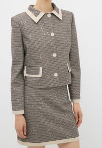 Brązowy garnitur w kratę houndstooth z krótką marynarką mającą kremowy kołnierz i guziki, w komplecie ze spódnicą i przednimi kieszeniami.