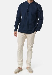 Chemise manches longues bleu marine avec col arrondi et patte de boutonnage à trois boutons, associée à un pantalon beige clair coupe slim et des chaussures grises sans lacets.