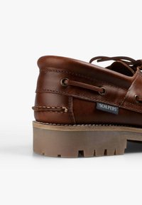 Zapato náutico de cuero marrón con un cuello redondeado, borde cosido y una suela de goma robusta. Cuenta con una etiqueta con logo y ojales metálicos para los cordones.