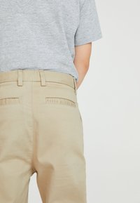 Bežové kalhoty s hladkým povrchem, s zadními kapsami a vyšitým štítkem s nápisem "BAKER BY TED BAKER." Kombinované s šedou košilí.
