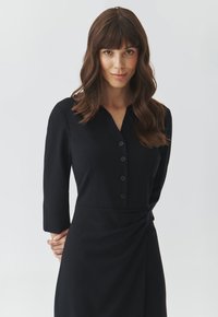 Schwarzes Kleid mit einem kragenlosen Ausschnitt, Knopfleiste und Dreiviertelärmeln. Verfügt über ein seitliches Twist-Detail und eine glatte Stoffqualität.