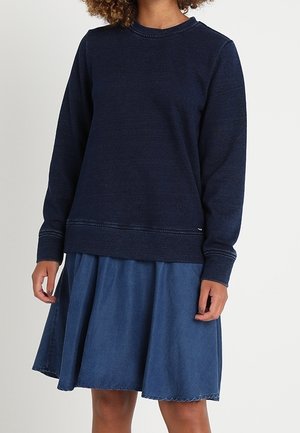 Woman wearing a navy blue long-sleeve sweatshirt and a knee-length blue denim skirt, standing against a plain white background.