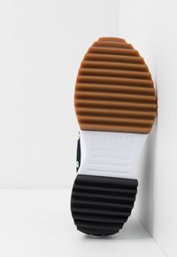 Svart och vit sneakersula med ribbad brun gummiyta, med en vit mellansula och varumärkesnamn präglat på hälen.