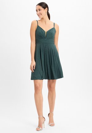Korte groene jurk met spaghettibandjes, glittertextuur, aansluitende bodice, geplooide rok en een diepe V-halslijn. Zilveren schoenen.