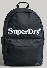 Zaino in tessuto nero con forma arrotondata, logo "SuperDry" bianco ben in evidenza, tasca frontale con zip e manico superiore per il trasporto.