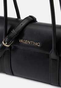 Černá kožená kabelka s zlatým nápisem "Valentino". Má nastavitelný černý popruh, hladký povrch a zaoblený tvar.