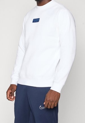 Mężczyzna w białej bluzie z niebieską naszywką "PARIS SAINT-GERMAIN" i granatowych spodniach dresowych Nike z białym logo Swoosh.