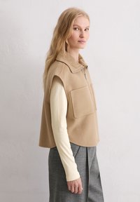 Beige fleecevest met een hoge kraag, ritssluiting aan de voorkant en twee grote opgestikte zakken, gedragen over een lichtgekleurde longsleeve en grijze broek.