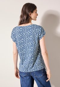 Top a maniche corte di colore blu scuro con motivo floreale blu chiaro. Vestibilità morbida, scollatura rotonda, tessuto soffice. Abbinato a jeans di denim blu.