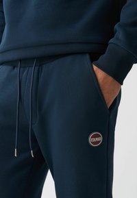 Mornarsko modri trening hlače z vrvico v pasu, stranskimi žepi, rebrastimi manšetami in okroglo oznako s "COLMAR" na stegnu.