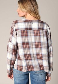 Geruite blouse met lange mouwen in witte, blauwe en bruine tinten; heeft een relaxte pasvorm, ronde halslijn en gathered mouwen. Knopdetail aan de achterkant.