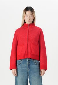 Jonge vrouw met lang steil haar die een helderrode trui met ritssluiting en voorkruiszakken draagt, samen met blauwe jeans, staande tegen een witte achtergrond.