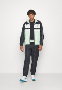 Sportinis treningas, kuriame yra mėtų žalios ir juodos spalvos užtrauktukas švarkas su baltais juostelėmis, derinamas su tamsiomis kelnėmis ir baltomis sportinėmis bateliais su spalvotais akcentais.