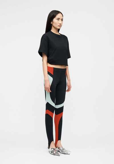 Νεαρή γυναίκα όρθια, με μαύρο κοντό T-shirt, μαύρο κολάν με κόκκινα και ανοιχτό μπλε γεωμετρικά σχέδια και μεταλλικά μυτερά παπούτσια.