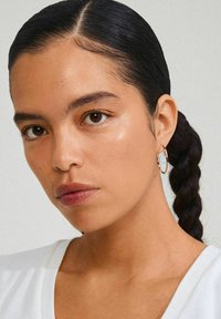 Pendientes de aro de oro con un sutil diseño texturizado, que presenta una mezcla de colores. El modelo tiene un peinado trenzado y lleva una prenda blanca.