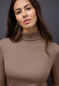 Bruin geribd coltruitje met een aansluitend model en een hoge kraag, gedragen door een vrouw met halflang haar tegen een donkere achtergrond.