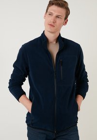Navy fleece jacket with a high collar, front zipper, and side pockets. Features a chest pocket with zip closing and ribbed cuffs.