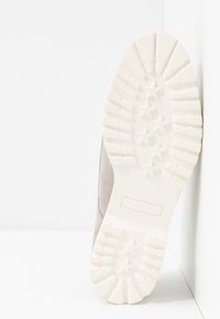 Semelle d'une chaussure beige avec des motifs de bande de roulement profonds incluant des formes d'étoiles, positionnée debout contre un mur d'angle blanc.