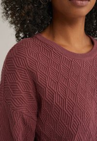 Pull en tricot texturé mauve avec un motif géométrique. Il possède un col rond côtelé et un tissu doux et épais.