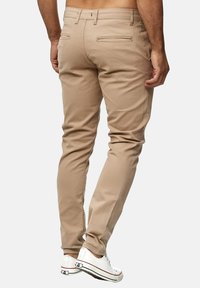 Beżowe chinosy o slim fit, z tylnymi kieszeniami, minimalnym przeszyciem i przetartymi mankietami, w zestawieniu z białymi trampkami.