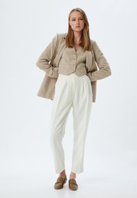 Blazer beige sopra un top corto abbinato con bottoni, abbinato a pantaloni bianchi plissettati e mocassini marroni. Texture lisce, vestibilità sartoriale.