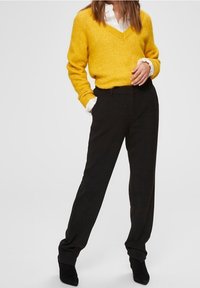 Jasnożółty sweter w serek, zestawiony z czarnymi spodniami na wysoki stan, o luźnym kroju i gładkim materiale. Teksturowane czarne botki za kostkę.